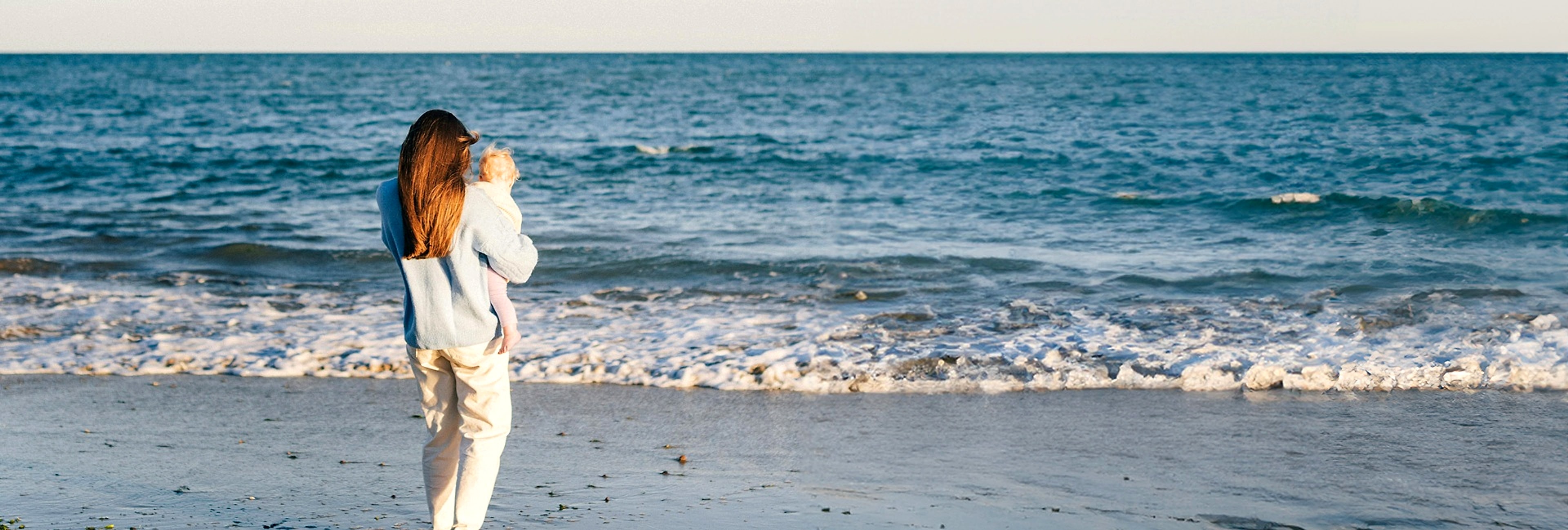 A mother holding her child at the shoreline, looking out to the horizon