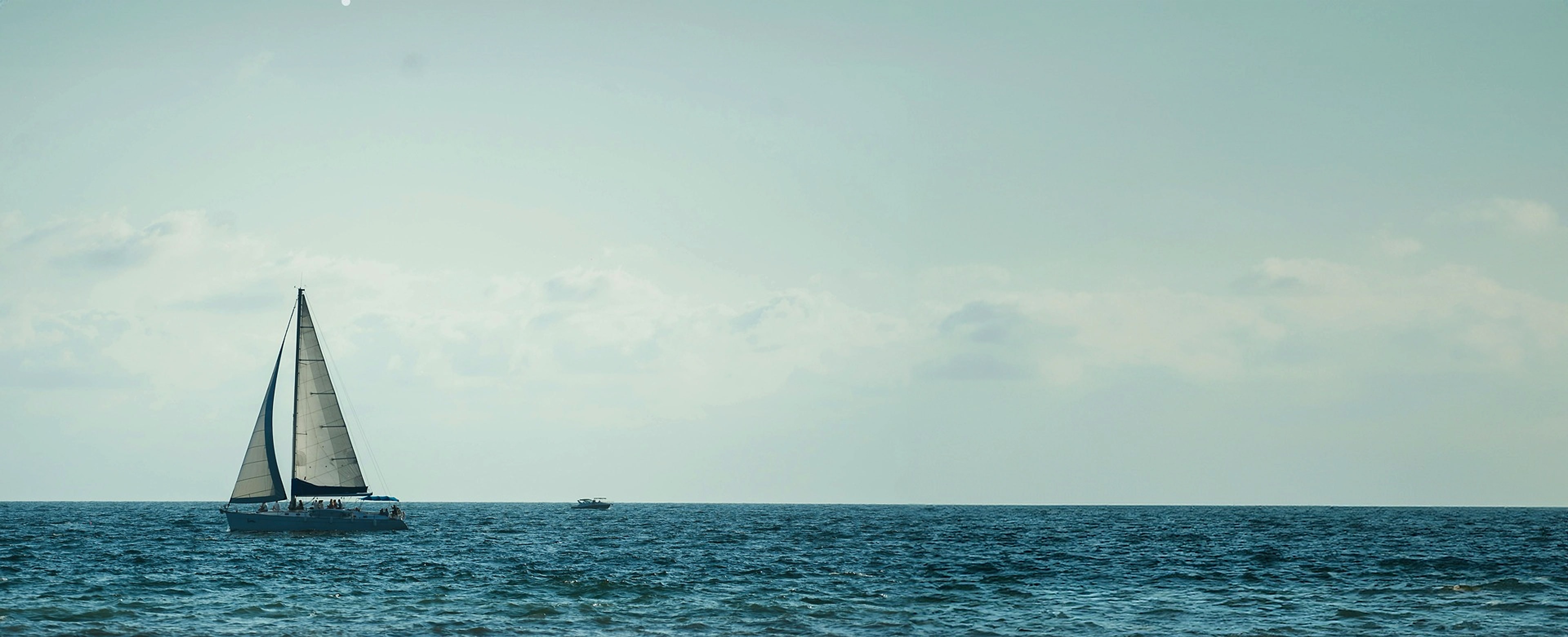 A sailboat crossing calm open water under a soft, bright sky